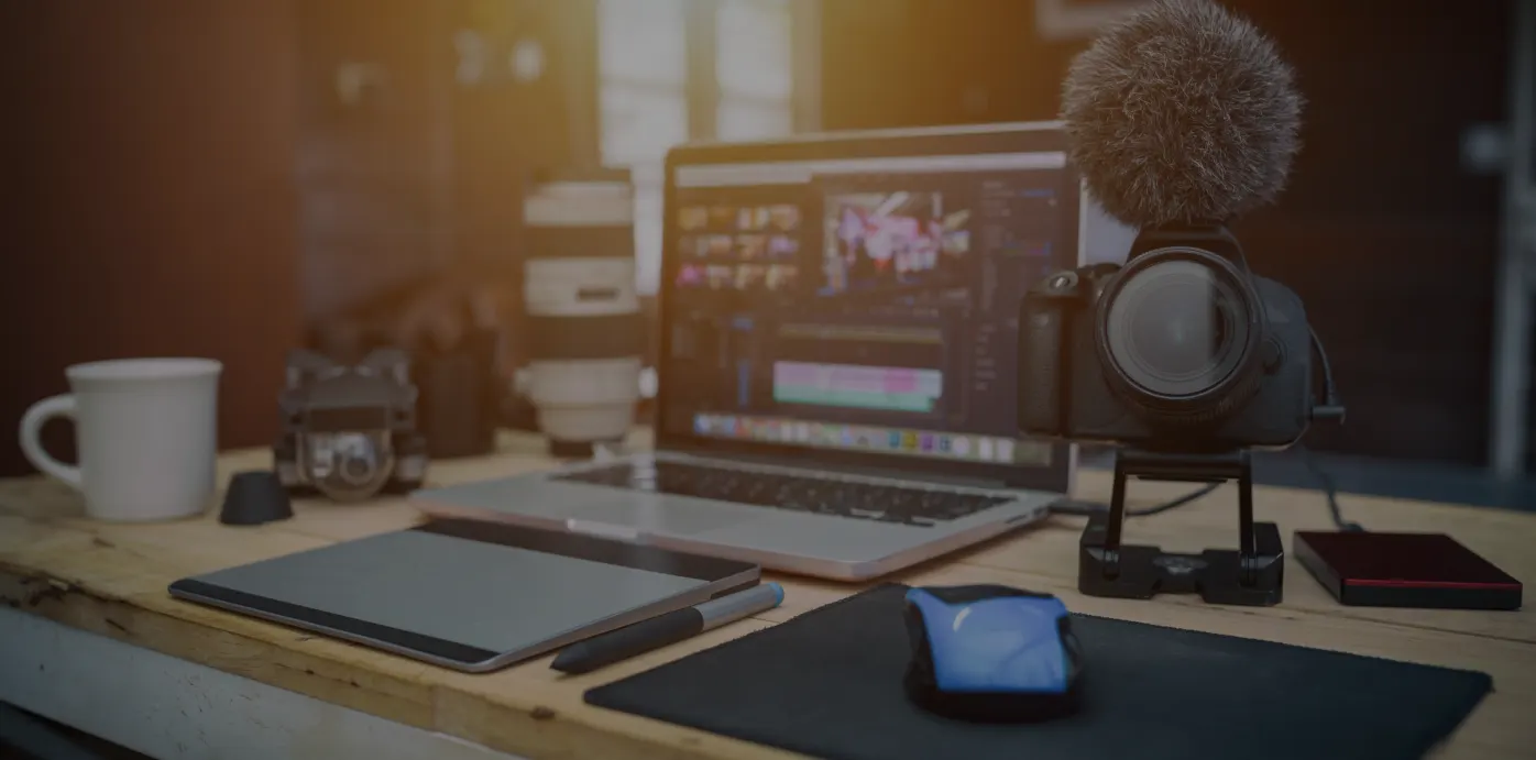 Workspace setup featuring a laptop running video editing software, professional camera with attached microphone, digital drawing pen, mouse, and coffee mug on a desk.